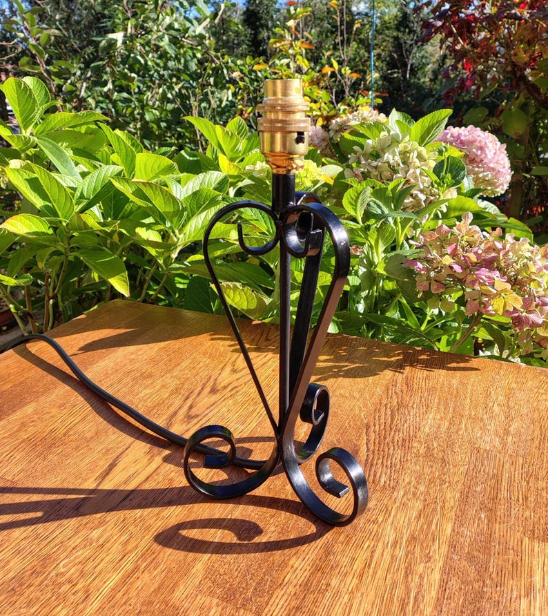 May include: Black wrought iron table lamp with a heart-shaped design. The lamp has a brass bulb socket and a decorative base with scrolled accents. The lamp is on a wooden surface, with greenery in the background.