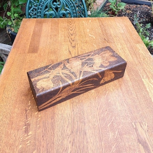 May include: A rectangular wooden box featuring a dark brown pyrography design of blooming flowers and long, slender leaves across the top surface. The box has a smooth, polished finish with visible wood grain patterns throughout the exterior.