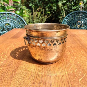 May include: A round brass planter with a weathered patina and a decorative band of heart-shaped cutouts near the rim. The vessel features two small side handles with attached rings and a smooth, rounded base, showing signs of age and oxidation.