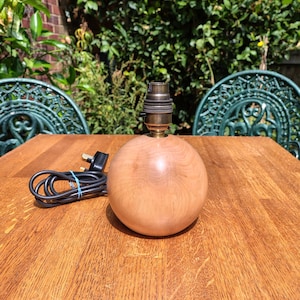 May include: A spherical light wood lamp base with a smooth, polished finish. The lamp features a metallic bronze-coloured bulb socket at the top and includes a black electrical cord with a standard plug attached to the side of the wooden sphere.