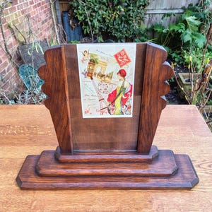 May include: A vintage wooden picture frame with an art deco design. The frame holds a print with Parisian imagery, including the Eiffel Tower and a woman in a red hat. The frame is made of dark wood and sits on a tiered base, perfect for a desk or shelf.