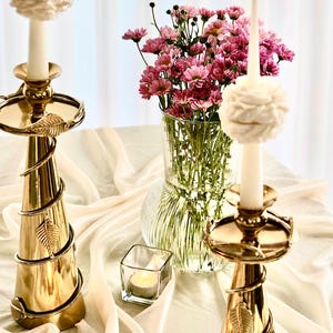 May include: Two gold candle holders with white candles and floral accents. The candles are lit and the holders are on a white tablecloth. A vase of pink flowers is in the background.