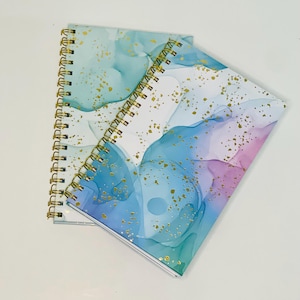 May include: Two spiral bound notebooks with a white background and blue and purple watercolor-like designs with gold glitter accents.
