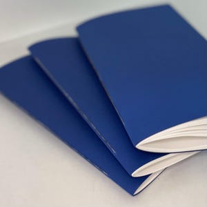 May include: Three blue notebooks stacked on top of each other. The notebooks have white pages and are bound with a white stitch.