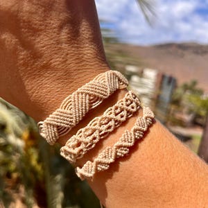Könnte beinhalten: Drei beige Makramee-Armbänder mit komplexen Knotenmustern. Das oberste Armband zeigt ein Chevron-Design, das mittlere Armband hat ein sich wiederholendes Muster aus offenen Schlaufen und das untere Armband weist eine kleine, enge Zickzack-Knotenstruktur auf.