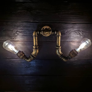 May include: A wall-mounted light fixture made from brass-colored pipes, featuring two exposed light bulbs. The fixture is mounted on a dark wooden surface. The fixture has a vintage, industrial aesthetic.