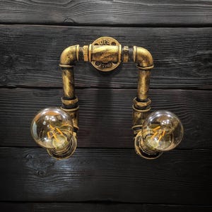 May include: A vintage-style wall sconce with a gold finish, featuring two exposed light bulbs. The fixture is constructed from pipes and fittings, with the text "UPS 1940" on the central plate. The sconce is mounted on a dark wood panel.