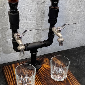 Può includere: Un distributore di liquori rustico con due beccucci, realizzato con tubi neri e rubinetti argentati, montato su una base di legno macchiato. Due bicchieri di cristallo sono posti sotto. Il distributore contiene due bottiglie di liquore ambrato.