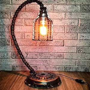 May include: A unique industrial-style table lamp. The lamp features a base made from a repurposed metal component, a curved chain arm, and a cage-like shade. The warm light from the bulb contrasts with the dark metal. The lamp is plugged into a wall outlet.