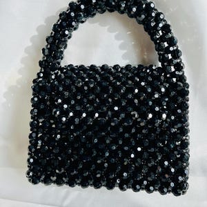 Puede incluir: Un bolso estructurado construido completamente con cuentas negras facetadas y brillantes. El bolso presenta un cuerpo rectangular y un asa superior redondeada, con todos los componentes unidos para formar una textura uniforme en forma de rejilla.