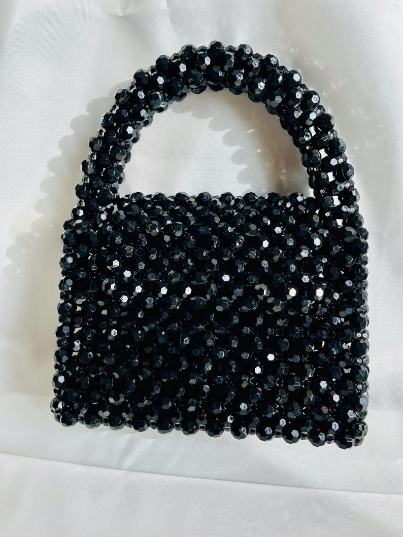 Puede incluir: Un bolso estructurado construido completamente con cuentas negras facetadas y brillantes. El bolso presenta un cuerpo rectangular y un asa superior redondeada, con todos los componentes unidos para formar una textura uniforme en forma de rejilla.