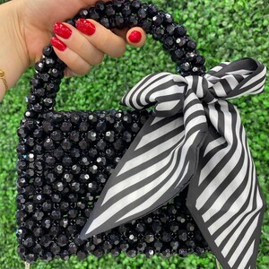Puede incluir: Un bolso peque&ntilde;o construido con cuentas negras facetadas, con un asa superior redondeada y un lazo decorativo de tela a rayas blancas y negras atado a un lado. El bolso incluye una fina correa de cadena de metal plateado en la base.