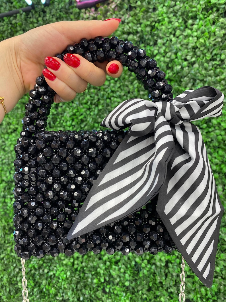 Puede incluir: Un bolso peque&ntilde;o construido con cuentas negras facetadas, con un asa superior redondeada y un lazo decorativo de tela a rayas blancas y negras atado a un lado. El bolso incluye una fina correa de cadena de metal plateado en la base.