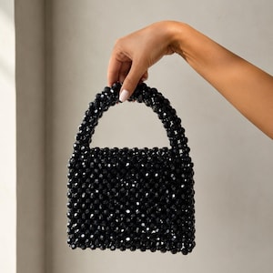 Puede incluir: Un bolso de mano peque&ntilde;o y cuadrado construido completamente con cuentas negras facetadas y brillantes. El bolso cuenta con un asa redondeada integrada, tambi&eacute;n hecha de las mismas cuentas negras, creando una apariencia texturizada y uniforme.