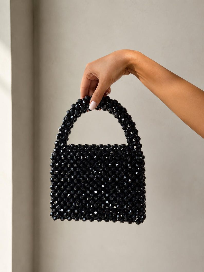 Puede incluir: Un bolso de mano peque&ntilde;o y cuadrado construido completamente con cuentas negras facetadas y brillantes. El bolso cuenta con un asa redondeada integrada, tambi&eacute;n hecha de las mismas cuentas negras, creando una apariencia texturizada y uniforme.