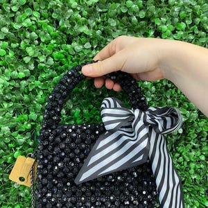 Puede incluir: Un bolso peque&ntilde;o cubierto de cuentas negras facetadas con un asa de cuentas a juego. Una cinta de tela a rayas blancas y negras est&aacute; atada en un lazo alrededor del asa. Una correa de cadena de metal fina y una peque&ntilde;a etiqueta beige est&aacute;n unidas.