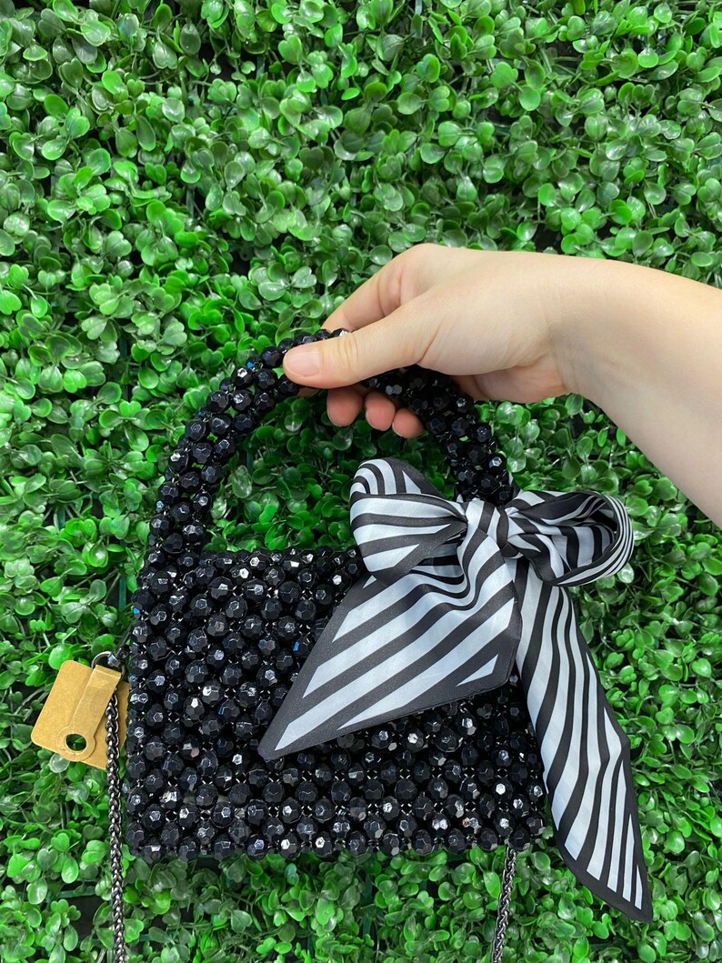 Puede incluir: Un bolso peque&ntilde;o cubierto de cuentas negras facetadas con un asa de cuentas a juego. Una cinta de tela a rayas blancas y negras est&aacute; atada en un lazo alrededor del asa. Una correa de cadena de metal fina y una peque&ntilde;a etiqueta beige est&aacute;n unidas.