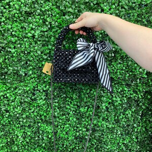 Puede incluir: Un bolso de mano negro hecho de cuentas con un cuerpo rectangular estructurado, un asa superior y una correa de cadena larga y fina. El bolso est&aacute; adornado con una cinta de tela a rayas blancas y negras atada en la parte delantera y una etiqueta.