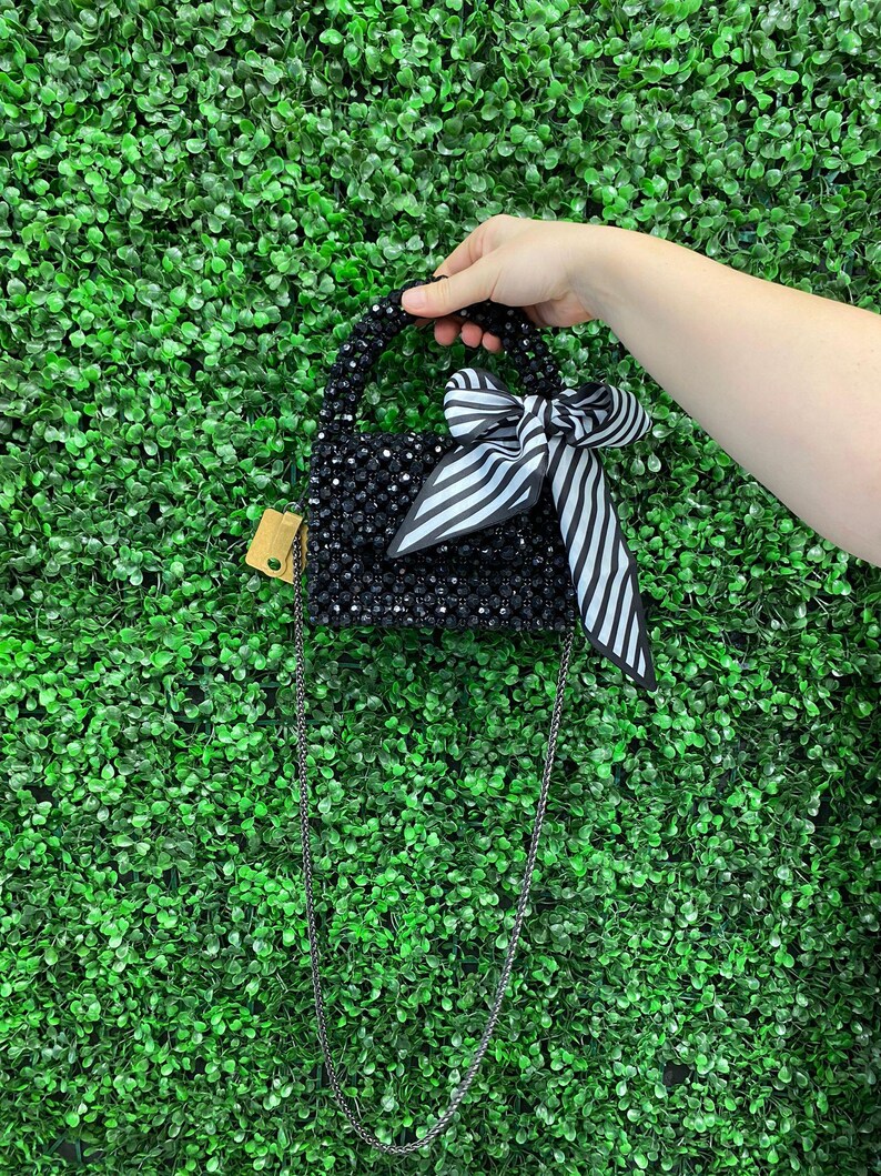 Puede incluir: Un bolso de mano negro hecho de cuentas con un cuerpo rectangular estructurado, un asa superior y una correa de cadena larga y fina. El bolso est&aacute; adornado con una cinta de tela a rayas blancas y negras atada en la parte delantera y una etiqueta.