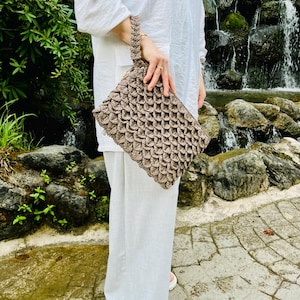 May include: A brown crocheted clutch bag with a scale-like pattern. The bag has a matching crocheted handle and is held in front of a person wearing white clothing. The background features a waterfall and greenery.