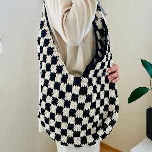 Może przedstawiać: Czarno-biała torba na ramię z wzorem szachownicy. Torba ma duży, zaokrąglony kształt z długim paskiem. Torba jest wykonana z miękkiej, teksturowanej przędzy. Torba jest trzymana przez osobę.