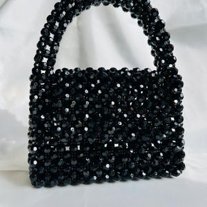 Puede incluir: Un bolso estructurado hecho completamente de peque&ntilde;as cuentas negras facetadas. El bolso presenta un cuerpo rectangular y un asa superior redondeada, ambos construidos con un patr&oacute;n de rejilla de cuentas negras reflectantes.