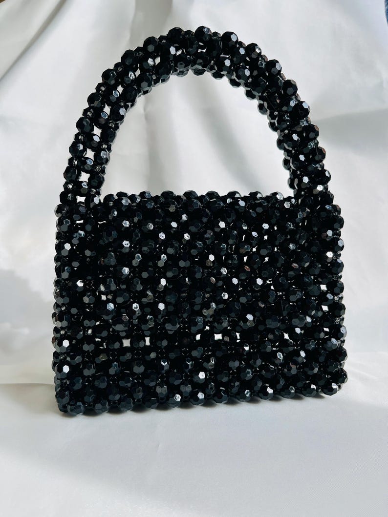 Puede incluir: Un bolso estructurado hecho completamente de peque&ntilde;as cuentas negras facetadas. El bolso presenta un cuerpo rectangular y un asa superior redondeada, ambos construidos con un patr&oacute;n de rejilla de cuentas negras reflectantes.