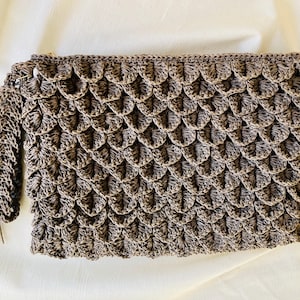 May include: A handmade, brown crochet clutch with a scale-like pattern. The clutch features a matching tassel and a wrist strap. The neutral color and textured design make it a versatile accessory for various outfits.