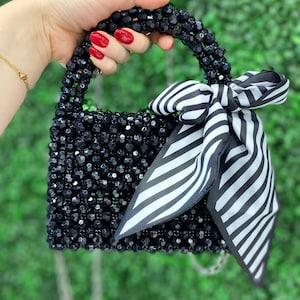 Puede incluir: Un bolso peque&ntilde;o construido con cuentas negras facetadas y un asa superior de cuentas a juego. Un pa&ntilde;uelo de tela con rayas blancas y negras est&aacute; atado en forma de lazo alrededor del asa. El bolso presenta un cuerpo rectangular y una fina cadena plateada.