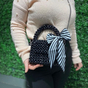 Puede incluir: Un bolso de mano peque&ntilde;o cubierto de cuentas facetadas con un asa de cuentas a juego. El bolso presenta un gran lazo decorativo hecho de tela a rayas blancas y negras sujeto a un lado. Incluye una fina correa de hombro de cadena de metal oscuro.