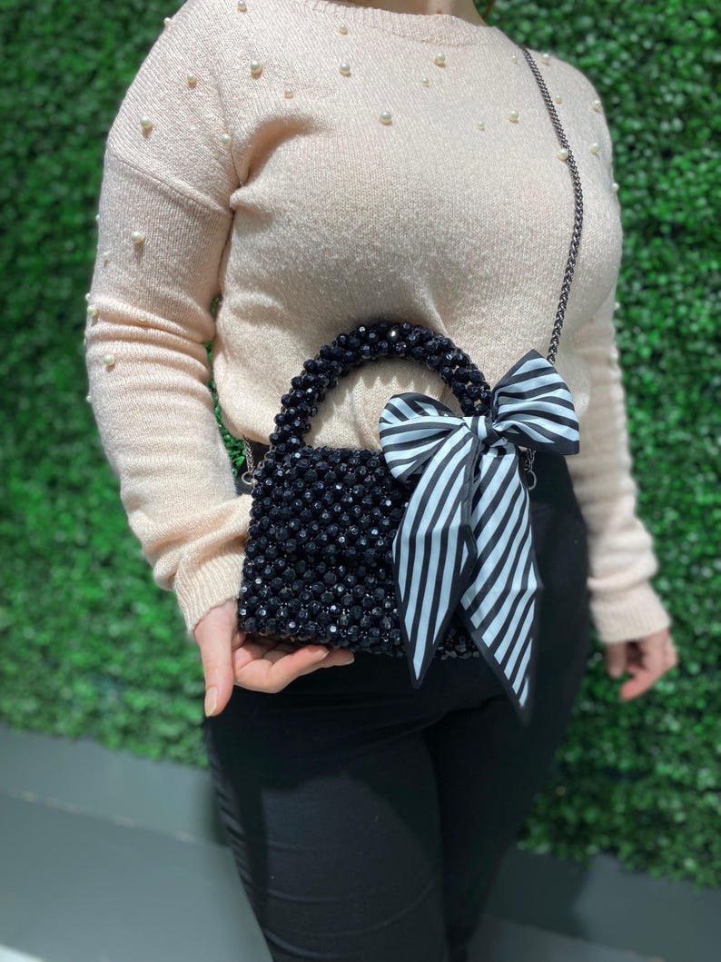 Puede incluir: Un bolso de mano peque&ntilde;o cubierto de cuentas facetadas con un asa de cuentas a juego. El bolso presenta un gran lazo decorativo hecho de tela a rayas blancas y negras sujeto a un lado. Incluye una fina correa de hombro de cadena de metal oscuro.