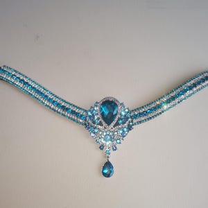 May include: A decorative browband featuring a black strap and a central jewel design. The jewel is a large, teal-coloured, teardrop-shaped stone, surrounded by smaller teal and clear crystals. The band is decorated with teal and clear crystals.