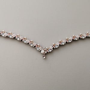 May include: A delicate, V-shaped bridal headpiece with a floral design. The headpiece is adorned with white and pink flowers, rhinestones, and pearls. A single rhinestone dangles from the centre of the piece.