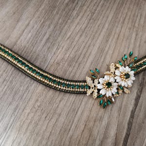 May include: A black leather horse browband with a gold and emerald green crystal design. The browband features a large, ornate floral design with white and green crystals.