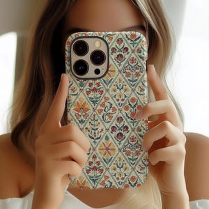 May include: A patterned phone case featuring a floral design in shades of red, orange, and blue on a cream background. The case has a diamond pattern with flowers and leaves. The phone case is held up in front of a person's face.