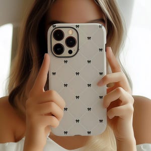 May include: A white phone case with a black bow pattern. The case has a grid design and a black camera lens border. The phone case is held up in front of a person's face.