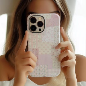 May include: A pink and white patchwork phone case with a floral and bow pattern. The case has a rectangular shape with rounded corners and a camera cutout. The design incorporates various textures and patterns, creating a visually appealing aesthetic.