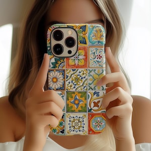 May include: A colorful phone case featuring a mosaic tile design with floral and geometric patterns in yellow, blue, orange, and green. The case has a camera cutout and is held in front of a person's face.