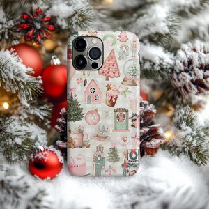 May include: A smartphone case featuring a repeating pattern of Christmas-themed illustrations including pink gingerbread houses, nutcrackers, Santa Claus, candy canes, decorated trees, snow globes, and mugs on a light green and white gingham background.