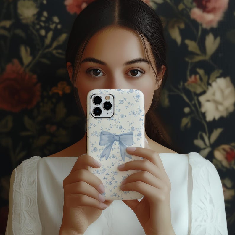 May include: A white phone case with a blue floral pattern and a blue bow design. The case is held in front of a person's face, with a floral patterned background.