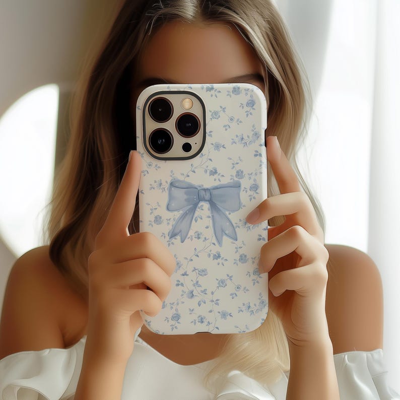 May include: A white phone case with a blue floral pattern and a light blue bow design. The case has a camera cutout and is held up in front of a person's face.