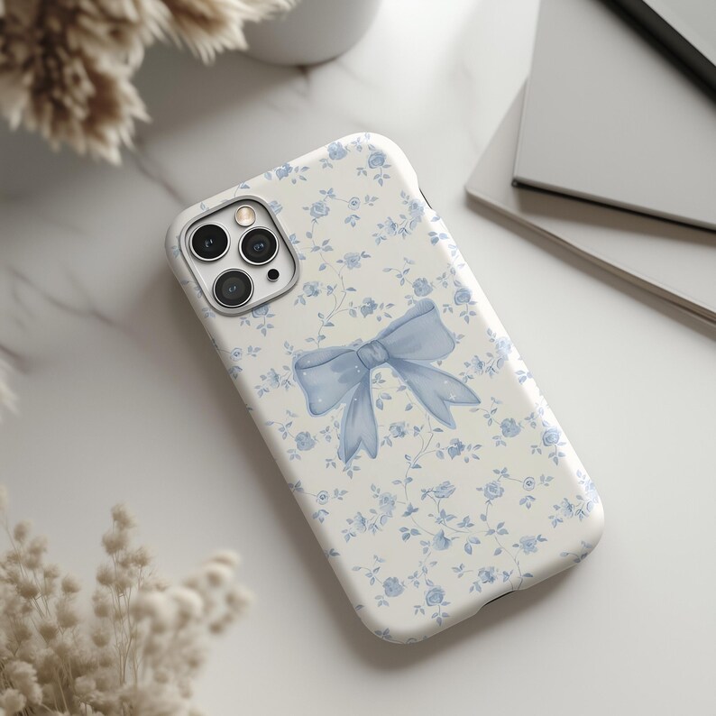 May include: A white phone case with a blue floral pattern and a light blue bow design. The case has a camera cutout and is lying on a white surface. The floral pattern includes small blue flowers and green stems.