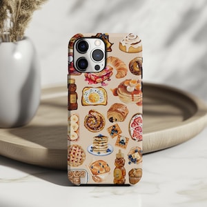 May include: A phone case featuring a watercolor illustration of various breakfast foods. The design includes pancakes, waffles, croissants, muffins, and other baked goods in a repeating pattern on a beige background.