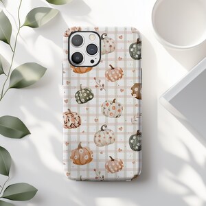 May include: A phone case featuring a repeating pattern of watercolor pumpkins in shades of orange, pink, and green. The pumpkins are decorated with floral and leaf designs, set against a beige and white checkered background with small heart accents.