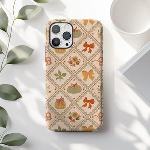 May include: A beige phone case with a repeating pattern of pumpkins, bows, sunflowers, and leaves. The design is arranged in a grid with a lace-like border. The case has a camera cutout and is on a white surface.