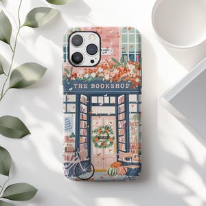 May include: A phone case featuring an illustrated bookshop scene. The design includes a pink building with a blue awning, a bicycle, pumpkins, and the text "THE BOOKSHOP". The case has a floral wreath on the door and a sign that reads "The world belongs to those who read".