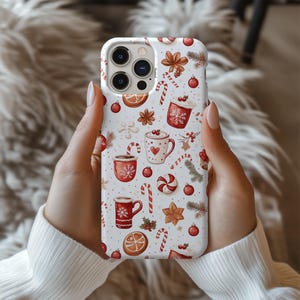 May include: A white phone case featuring a festive Christmas pattern. The design includes red mugs, candy canes, gingerbread cookies, and ornaments. The case has a glossy finish and is held by a person wearing a white sweater.