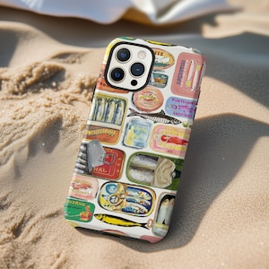 May include: A phone case featuring a colorful design of vintage sardine and anchovy tins. The case has a white background with various tin labels in shades of red, yellow, blue, and green. The phone case is lying on a sandy surface.