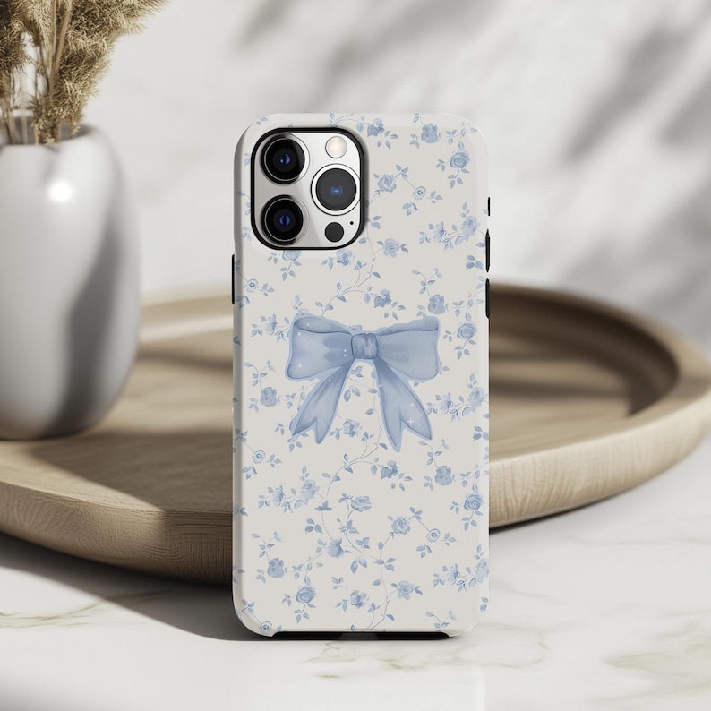 May include: A white phone case with a light blue bow and floral pattern. The case features a large bow in the center and small blue flowers scattered across the surface. The phone case is designed to fit a smartphone.