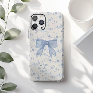 May include: A phone case featuring a light blue bow design. The case has a floral pattern with small blue flowers on a cream background. The phone case is a protective accessory for a smartphone.